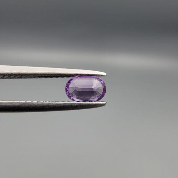 Studio close-up photograph displaying a 0.37-carat oval-cut natural amethyst Katela gemstone held securely in precision tweezers against a neutral grey background.