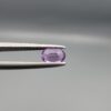 Studio close-up photograph displaying a 0.37-carat oval-cut natural amethyst Katela gemstone held securely in precision tweezers against a neutral grey background.