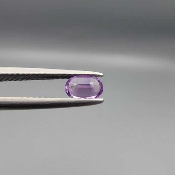 Studio close-up photograph displaying a 0.37-carat oval-cut natural amethyst Katela gemstone held securely in precision tweezers against a neutral grey background.