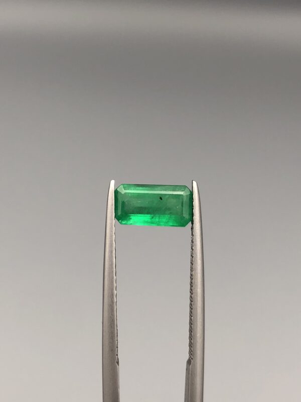 Studio close-up photographs showing the top-down view of an elongated rectangular step-cut 1.21 carat natural green emerald gemstone held securely in precision tweezers.
