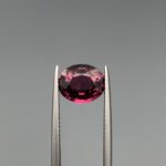 ​A 2.78-carat oval faceted natural purplish-pink garnet held in metal tweezers against a neutral gray background, displaying vibrant magenta flashes and high transparency.