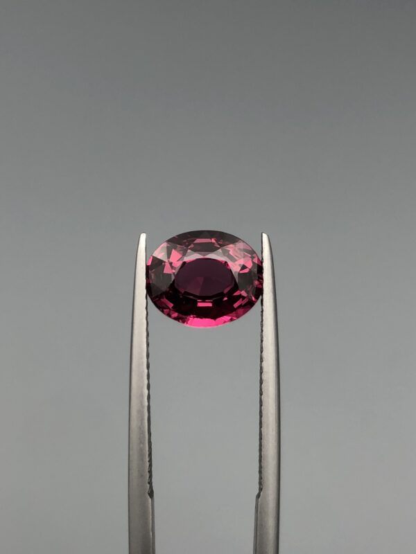 ​A 2.78-carat oval faceted natural purplish-pink garnet held in metal tweezers against a neutral gray background, displaying vibrant magenta flashes and high transparency.