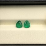 Studio close-up photographs displaying a matched pair of pear-shaped natural green emerald gemstones resting side-by-side inside an open black presentation box.