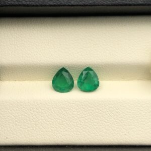 Studio close-up photographs displaying a matched pair of pear-shaped natural green emerald gemstones resting side-by-side inside an open black presentation box.