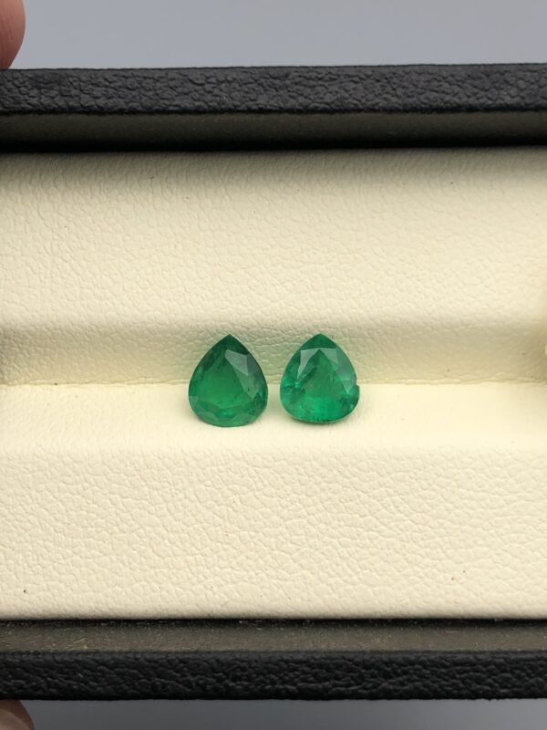 Studio close-up photographs displaying a matched pair of pear-shaped natural green emerald gemstones resting side-by-side inside an open black presentation box.