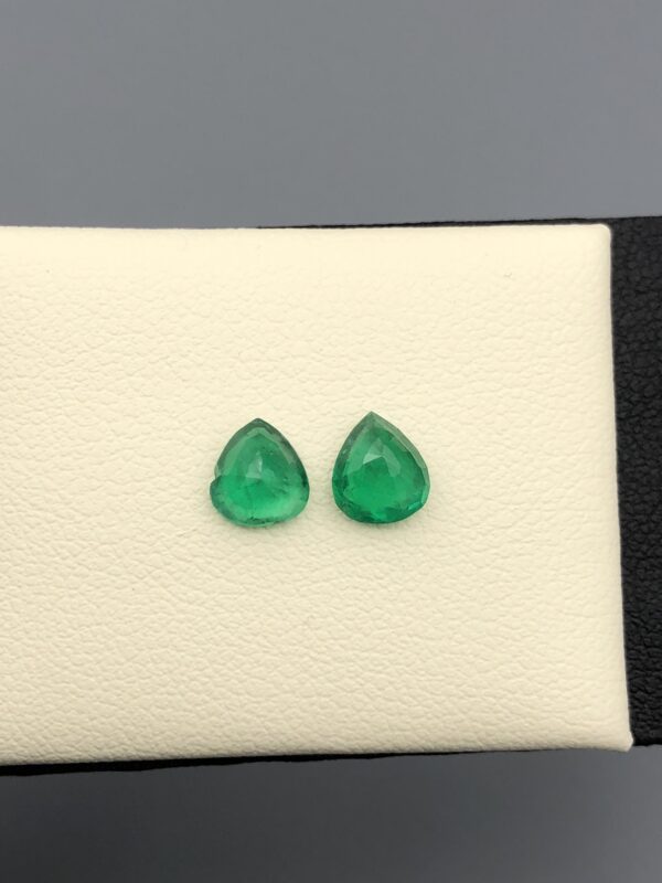 Studio close-up photographs displaying a matched pair of pear-shaped natural green emerald gemstones resting side-by-side inside an open black presentation box.
