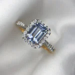 Close-up photograph of an 18K gold ring featuring a 1.53-carat emerald-cut pastel blue natural Ceylon sapphire centerpiece surrounded by a shimmering halo of 0.33-carat natural diamond accents against a soft white fabric background.