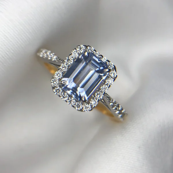 Close-up photograph of an 18K gold ring featuring a 1.53-carat emerald-cut pastel blue natural Ceylon sapphire centerpiece surrounded by a shimmering halo of 0.33-carat natural diamond accents against a soft white fabric background.