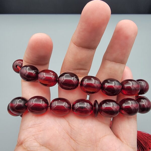 Studio close-up photograph of a 60-gram deep red German Bakelite Sandaloos tasbih rosary, featuring barrel-cut polished beads, an intricately sculpted imam bead, and a rich red cord tassel against a light background.