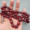 Studio close-up photograph of a 60-gram deep red German Bakelite Sandaloos tasbih rosary, featuring barrel-cut polished beads, an intricately sculpted imam bead, and a rich red cord tassel against a light background.