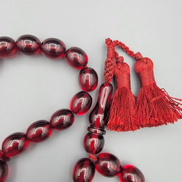 Studio close-up photograph of a 60-gram deep red German Bakelite Sandaloos tasbih rosary, featuring barrel-cut polished beads, an intricately sculpted imam bead, and a rich red cord tassel against a light background.
