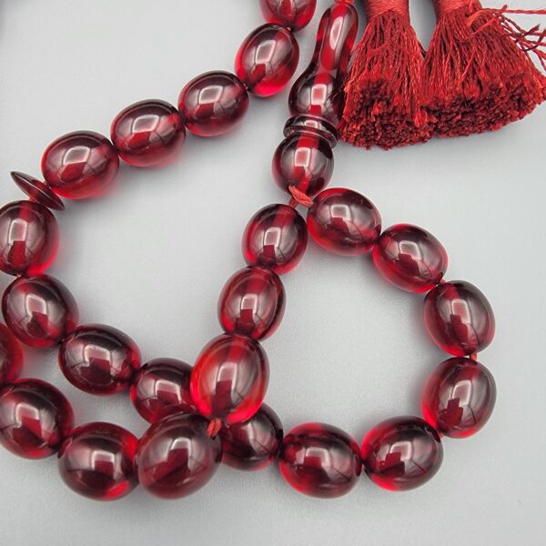 Studio close-up photograph of a 60-gram deep red German Bakelite Sandaloos tasbih rosary, featuring barrel-cut polished beads, an intricately sculpted imam bead, and a rich red cord tassel against a light background.
