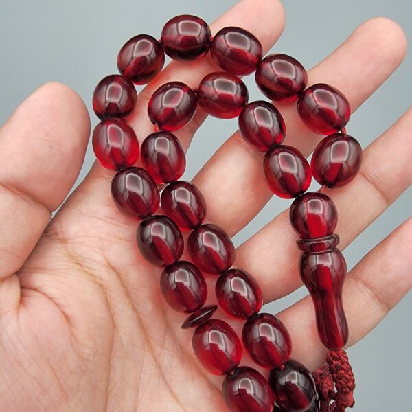 Studio close-up photograph of a 60-gram deep red German Bakelite Sandaloos tasbih rosary, featuring barrel-cut polished beads, an intricately sculpted imam bead, and a rich red cord tassel against a light background.
