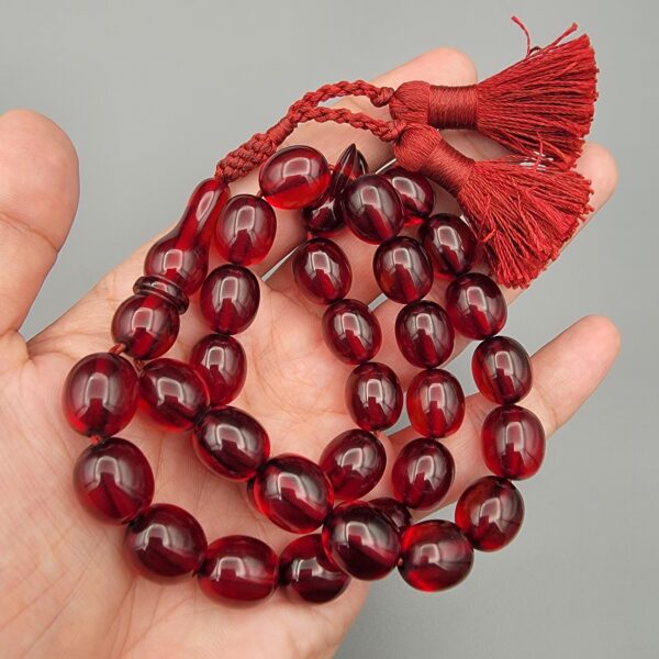 Studio close-up photograph of a 60-gram deep red German Bakelite Sandaloos tasbih rosary, featuring barrel-cut polished beads, an intricately sculpted imam bead, and a rich red cord tassel against a light background.