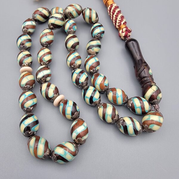 Studio close-up photograph of a 39-gram natural Yusr coral retro swirl striped composite tasbih prayer bead strand, featuring barrel-cut beads with turquoise-blue, maroon, and cream accents, a matching imam bead, and a multi-colored tassel against a light grey background.