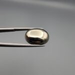 Studio close-up photographs displaying an 18.82-carat oval-cut natural brassy-gold pyrite gemstone with a metallic luster, held securely in precision tweezers against a neutral grey background.