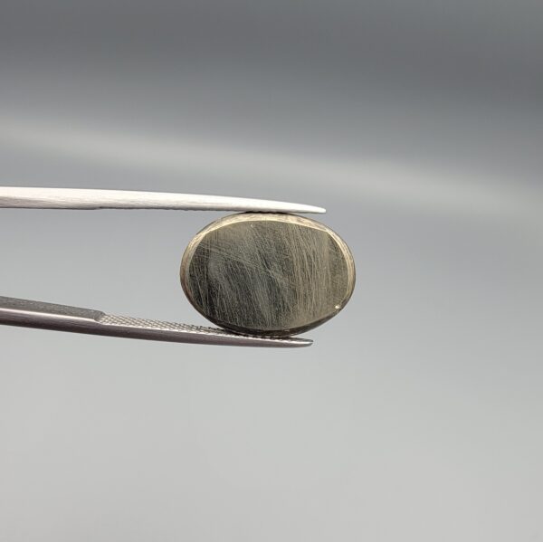 Studio close-up photographs displaying an 18.82-carat oval-cut natural brassy-gold pyrite gemstone with a metallic luster, held securely in precision tweezers against a neutral grey background.