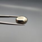 Studio close-up photographs showcasing a 16.38-carat oval-cut natural brassy-gold pyrite gemstone with a metallic luster, held securely in precision tweezers against a neutral grey background.