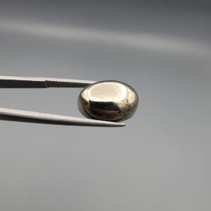 Studio close-up photographs showcasing a 16.38-carat oval-cut natural brassy-gold pyrite gemstone with a metallic luster, held securely in precision tweezers against a neutral grey background.