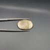 Studio close-up photographs showcasing a 16.38-carat oval-cut natural brassy-gold pyrite gemstone with a metallic luster, held securely in precision tweezers against a neutral grey background.