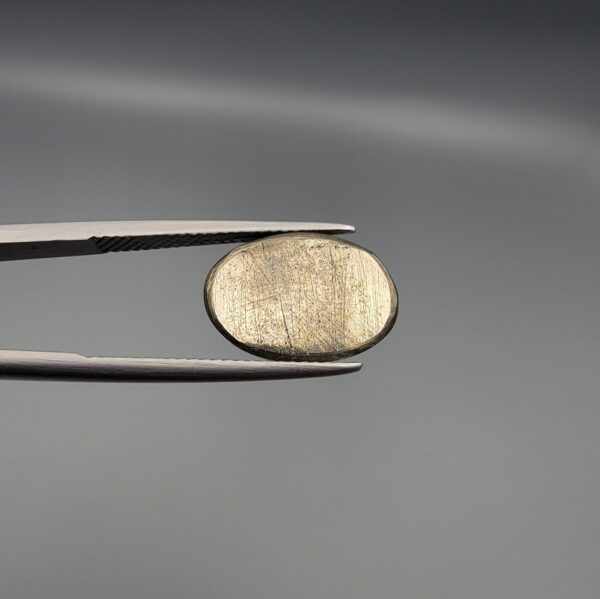 Studio close-up photographs showcasing a 16.38-carat oval-cut natural brassy-gold pyrite gemstone with a metallic luster, held securely in precision tweezers against a neutral grey background.