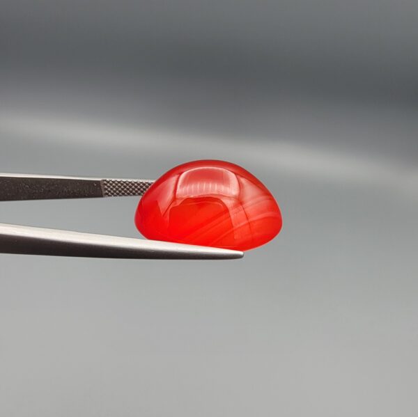 Studio close-up photographs displaying a 20.02-carat oval-cut natural radiant ember orangish-red agate Aqeeq gemstone, held securely in precision tweezers against a neutral grey background.