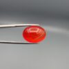 Studio close-up photographs displaying a 20.02-carat oval-cut natural radiant ember orangish-red agate Aqeeq gemstone, held securely in precision tweezers against a neutral grey background.