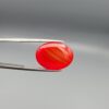 Studio close-up photographs displaying a 20.02-carat oval-cut natural radiant ember orangish-red agate Aqeeq gemstone, held securely in precision tweezers against a neutral grey background.