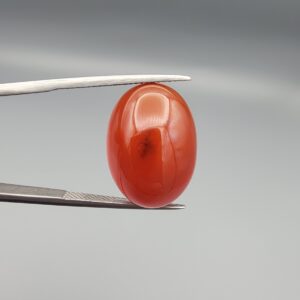 Studio close-up photograph showcasing a 22.63-carat natural carnelian agate Aqeeq cabochon with unique visible dark inclusions, held securely in precision tweezers against a neutral background.