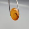 Studio close-up photograph displaying a 31.62-carat oval-cut natural honey-orange carnelian agate gemstone featuring a distinctive central dark inclusion pattern, held securely in precision tweezers against a neutral grey background.