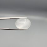 Studio close-up photograph displaying a 15.31-carat natural Durr-e-Najaf oval cabochon gemstone of spiritual purity held securely in precision tweezers against a neutral grey background.