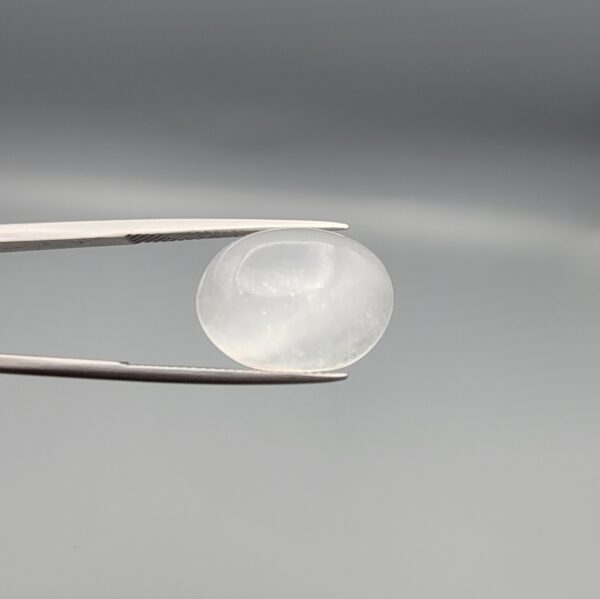 Studio close-up photograph displaying a 15.31-carat natural Durr-e-Najaf oval cabochon gemstone of spiritual purity held securely in precision tweezers against a neutral grey background.