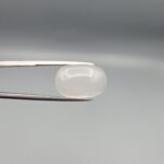 Studio close-up photograph displaying a 15.94-carat natural Durr-e-Najaf oval cabochon gemstone of spiritual purity held securely in precision tweezers against a neutral grey background.