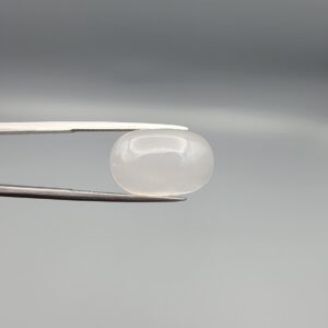Studio close-up photograph displaying a 15.94-carat natural Durr-e-Najaf oval cabochon gemstone of spiritual purity held securely in precision tweezers against a neutral grey background.