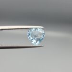 Studio close-up photograph displaying a 3.07-carat heart-cut natural sky blue topaz gemstone held securely in precision tweezers against a neutral grey background.