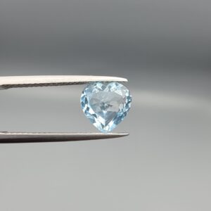 Studio close-up photograph displaying a 3.07-carat heart-cut natural sky blue topaz gemstone held securely in precision tweezers against a neutral grey background.