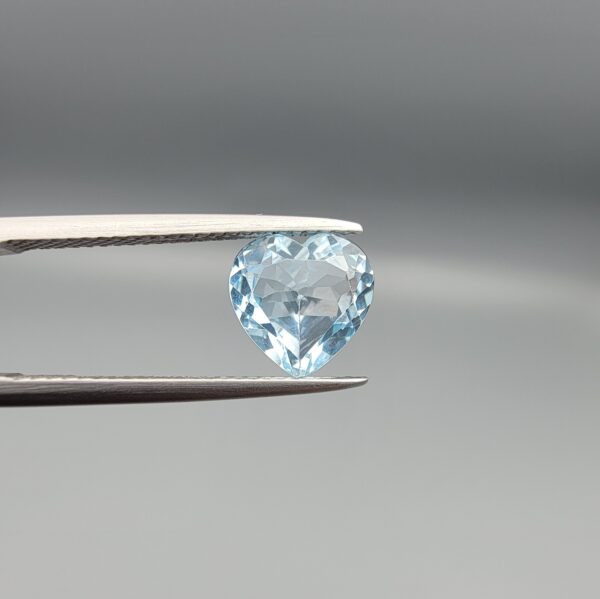 Studio close-up photograph displaying a 3.07-carat heart-cut natural sky blue topaz gemstone held securely in precision tweezers against a neutral grey background.