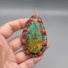 Hand holding a large, teardrop-shaped sterling silver statement pendant featuring a webbed desert matrix turquoise center stone surrounded by cabochon red coral stones and intricate filigree silverwork against a grey background.
