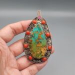 Hand holding a large, teardrop-shaped sterling silver statement pendant featuring a webbed desert matrix turquoise center stone surrounded by cabochon red coral stones and intricate filigree silverwork against a grey background.