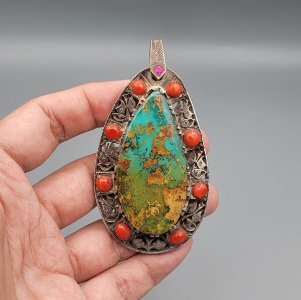 Hand holding a large, teardrop-shaped sterling silver statement pendant featuring a webbed desert matrix turquoise center stone surrounded by cabochon red coral stones and intricate filigree silverwork against a grey background.