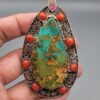 Hand holding a large, teardrop-shaped sterling silver statement pendant featuring a webbed desert matrix turquoise center stone surrounded by cabochon red coral stones and intricate filigree silverwork against a grey background.