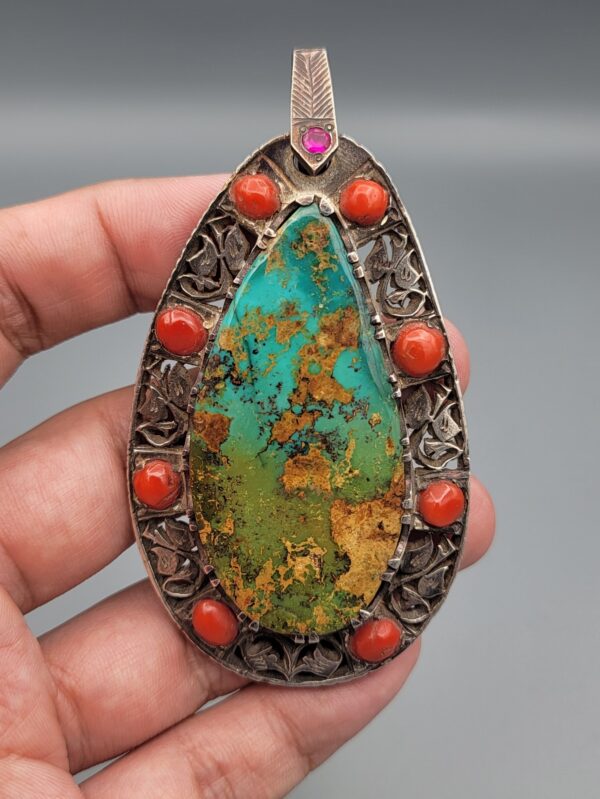 Hand holding a large, teardrop-shaped sterling silver statement pendant featuring a webbed desert matrix turquoise center stone surrounded by cabochon red coral stones and intricate filigree silverwork against a grey background.