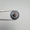 Close-up photograph of a 14.58-carat natural black eye agate cabochon gemstone held in stainless steel tweezers against a neutral grey background, displaying a striking concentric natural "eye" banding pattern.