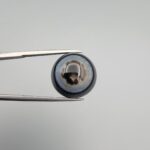 Close-up photograph of a 14.58-carat natural black eye agate cabochon gemstone held in stainless steel tweezers against a neutral grey background, displaying a striking concentric natural "eye" banding pattern.