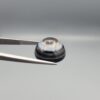 Close-up photograph of a 14.58-carat natural black eye agate cabochon gemstone held in stainless steel tweezers against a neutral grey background, displaying a striking concentric natural "eye" banding pattern.