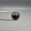 Close-up photograph of a 12.96-carat natural black eye agate cabochon gemstone held in stainless steel tweezers against a neutral grey background, displaying precise white concentric ring banding against a rich black body.