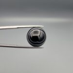 Close-up photograph of a 12.96-carat natural black eye agate cabochon gemstone held in stainless steel tweezers against a neutral grey background, displaying precise white concentric ring banding against a rich black body.