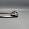 Close-up photograph of a 12.96-carat natural black eye agate cabochon gemstone held in stainless steel tweezers against a neutral grey background, displaying precise white concentric ring banding against a rich black body.
