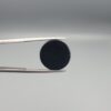 Close-up photograph of a 6.80-carat natural black eye agate cabochon gemstone held in stainless steel tweezers against a neutral grey background, featuring a distinct, sharp white circular eye banding pattern on a jet-black body.