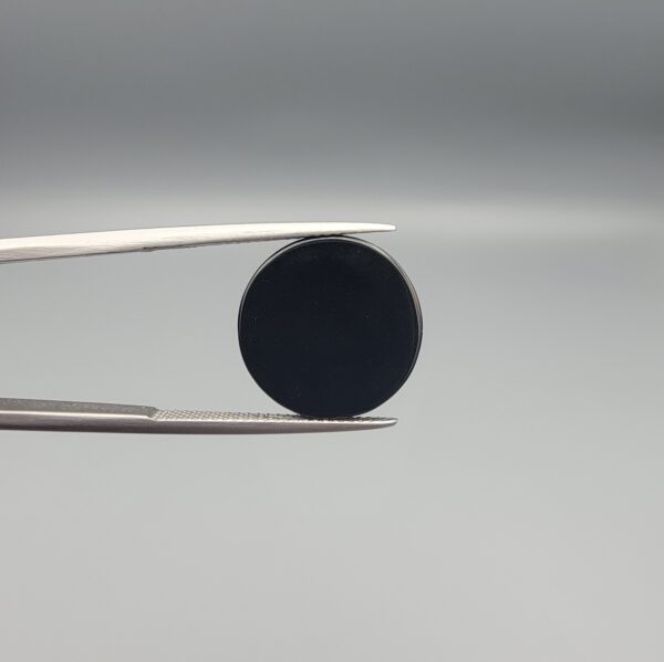 Close-up photograph of a 6.80-carat natural black eye agate cabochon gemstone held in stainless steel tweezers against a neutral grey background, featuring a distinct, sharp white circular eye banding pattern on a jet-black body.